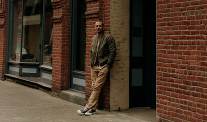 fit model leaning agains the wall wearing The Bomber Jacket in Fatigue Olive Dry Wax