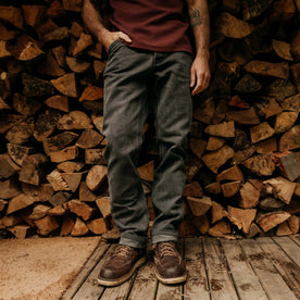 fit model leaning against a wood pile wearing The Camp Pant in Coal Chipped Canvas, Bottoms by Taylor Stitch