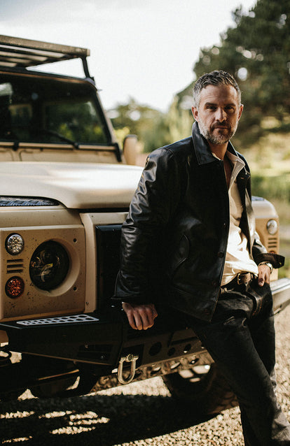 Sitting on the front bumper of a Landrover, hand ticked in pocket of jeans.