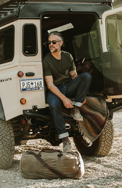 Sitting on the rear door, adjusting the roll on bottom of denim jeans, a duffel bag in the foreground.