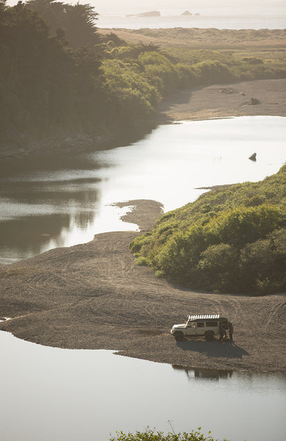 Aerial shot of a Landrover parked on a river bank, with two people looing in the back.