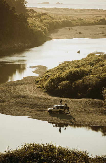 Two guys unloading duffel bags from a Landrover parked on a river bank.