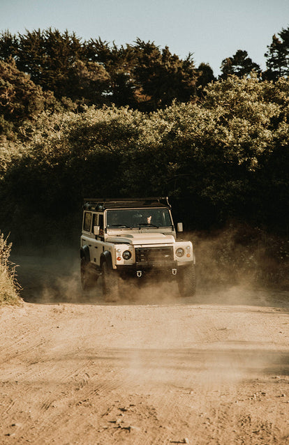 A Landrover kicking up dust as it travels down a tree-lined dirt road.