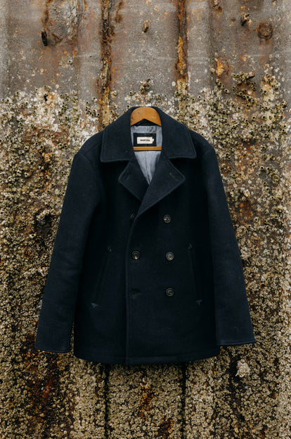 The Mariner Coat in Dark Navy hanging on a barnacle-covered wall