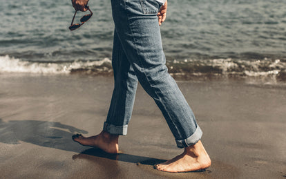 A guy's legs, walking barefoot down the beach in rolled up denim.